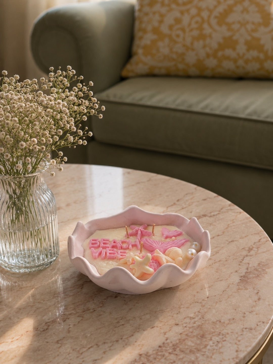 A scalloped pink ceramic dish containing pink and white decorative candles, styled on a round marble coffee table next to a glass vase of baby's breath in a living room setting.