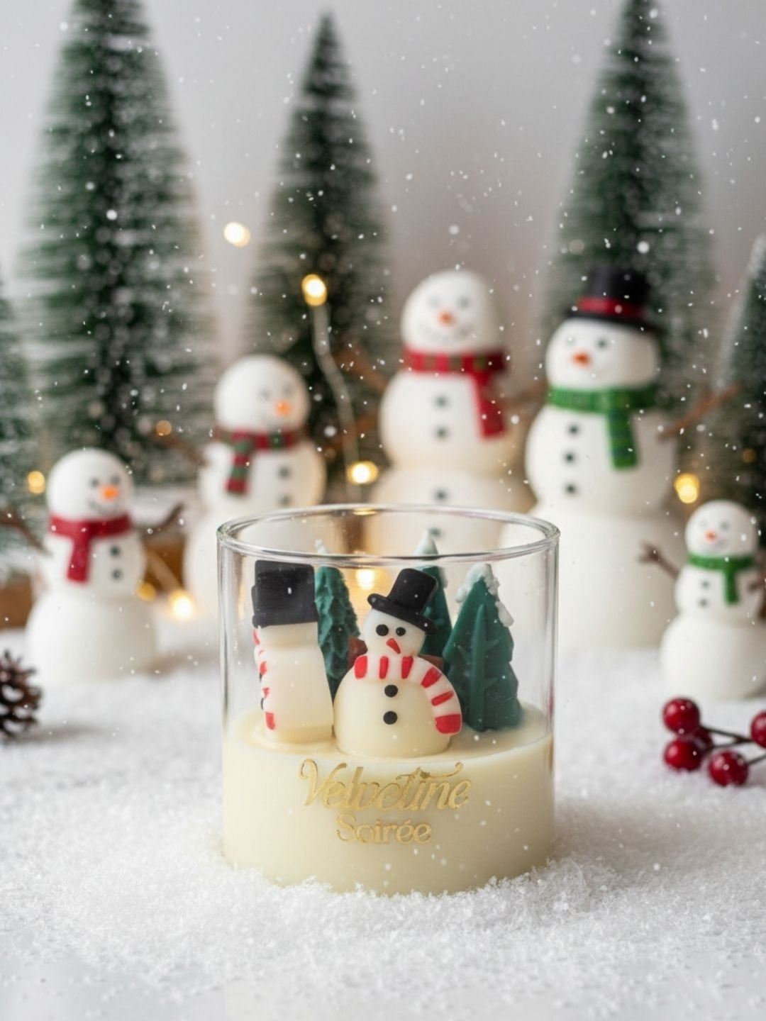 A clear glass jar candle filled with creamy off-white wax, decorated with miniature snowman figures and small green trees, set in a snowy display with blurred Christmas trees and snowman ornaments in the background.