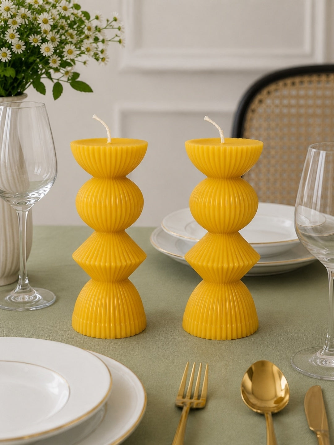 Two bright yellow ribbed pillar candles shaped like stacked cups, displayed on a styled dining table with white plates, gold cutlery, wine glasses, and a vase of small white flowers. Velvetine Soirée Beeswax Candle Pillar Candles