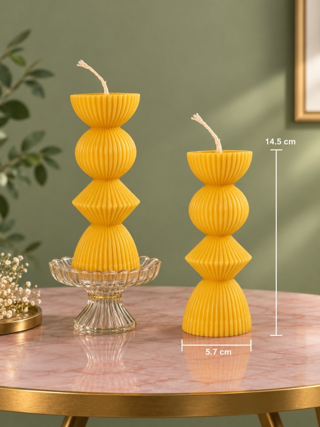 Two yellow ribbed pillar candles displayed on a pink marble table against a sage green wall, with dimension lines showing 14.5 cm height and 5.