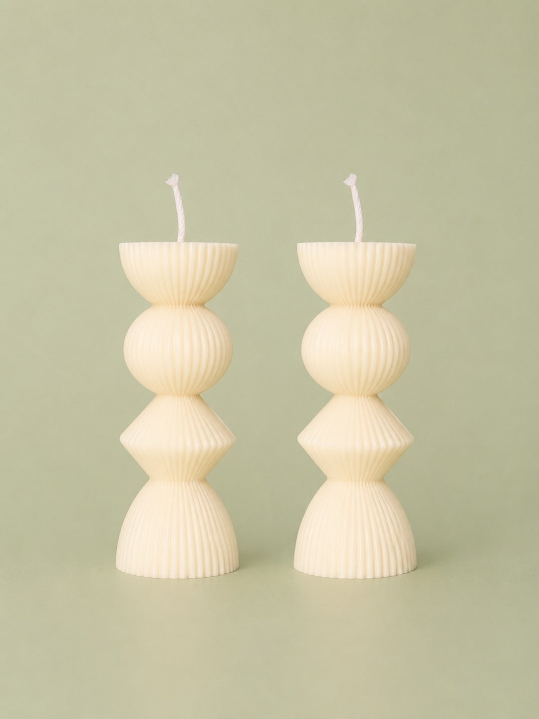 Two ribbed pillar candles in a cream color with a stacked, geometric design, standing on a matching light sage green background. Velvetine Soirée Beeswax Candle Pillar Candles