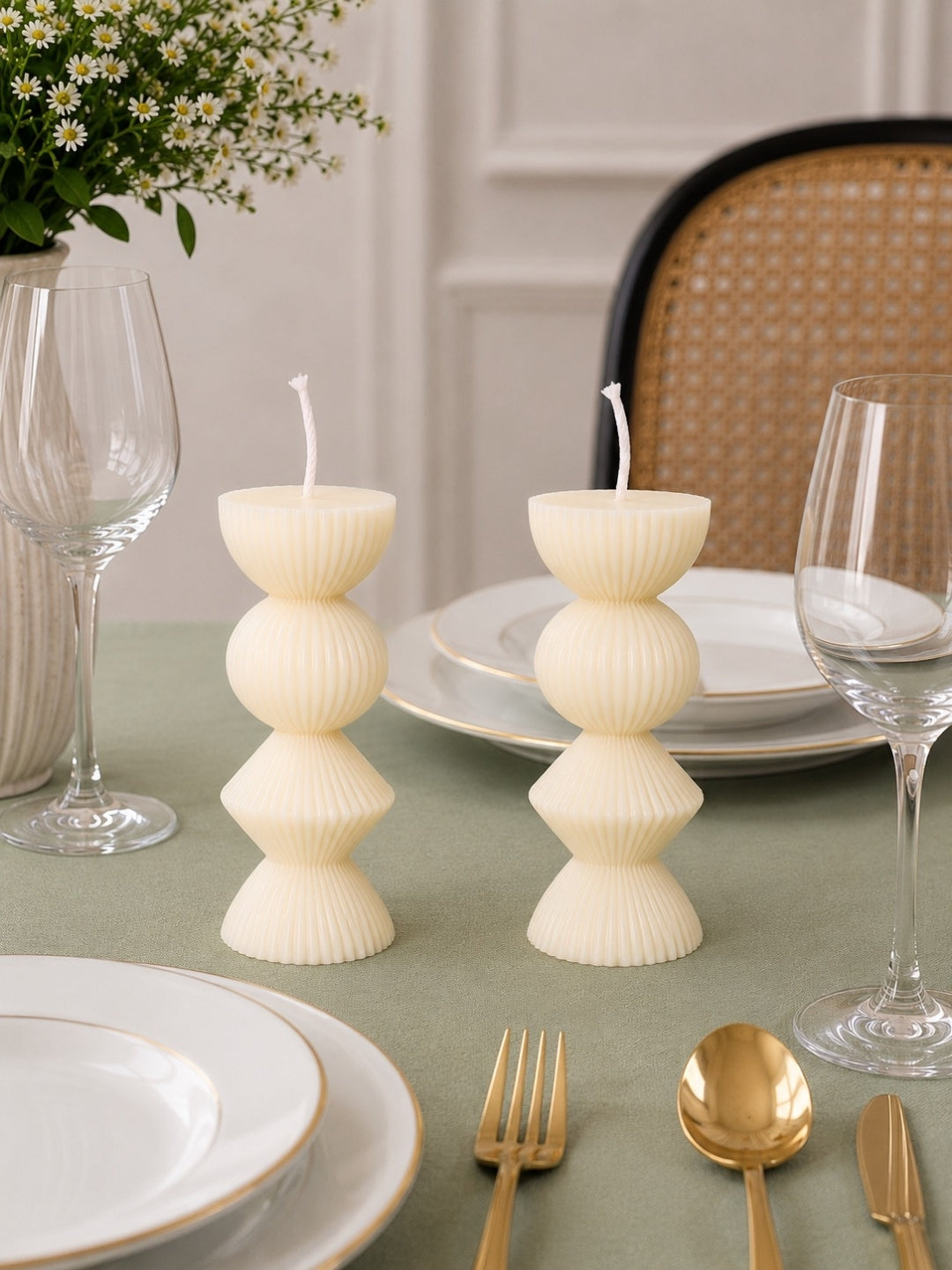 Two White-colored ribbed stacked pillar candles on a green tablecloth, styled with white plates, gold flatware, wine glasses, and a woven chair in the background.