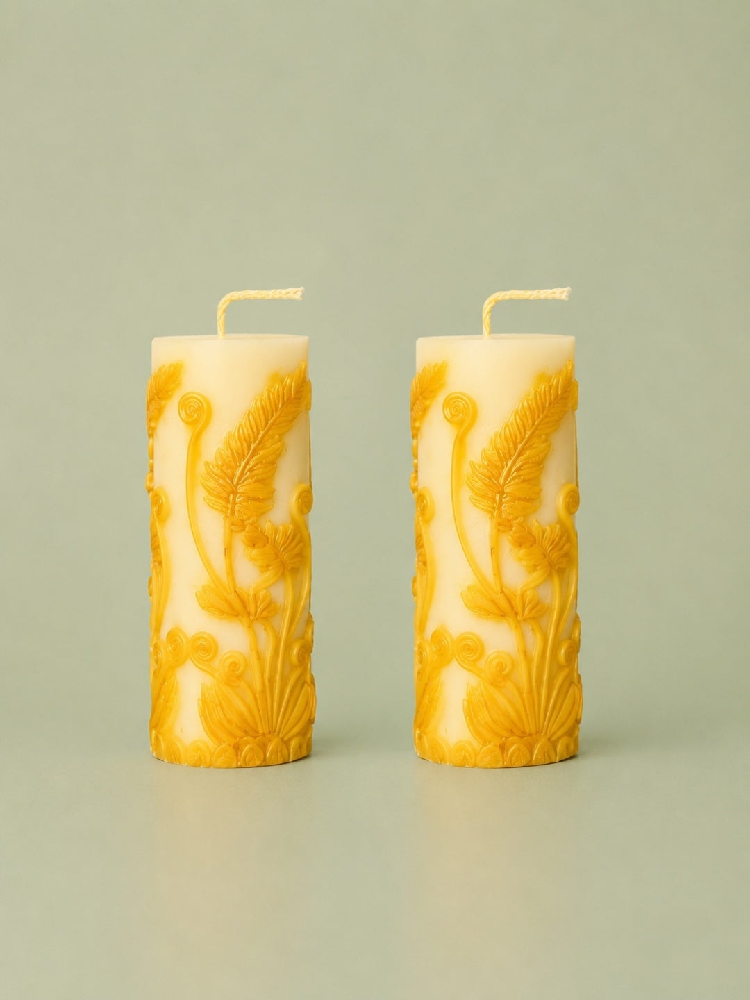 Two cream-colored pillar candles with yellow embossed floral scrollwork, standing upright on a light gray surface against a matching light gray background.