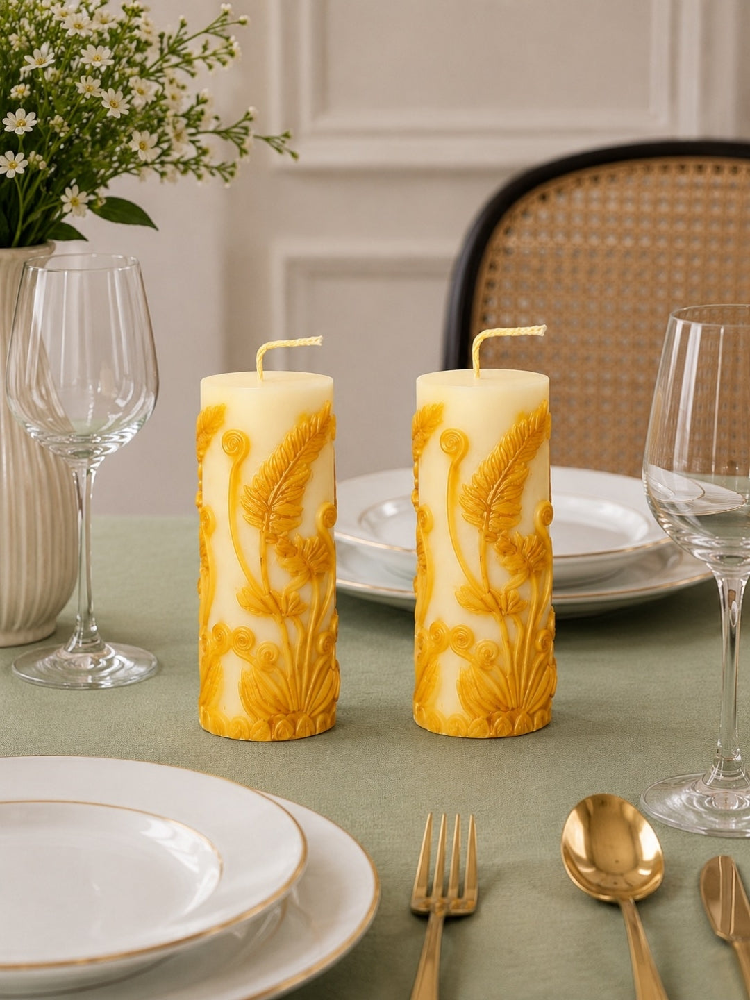 Two White pillar candles with raised floral and leaf patterns, displayed on a dining table with plates, wine glasses, gold cutlery, and a vase of white flowers in the background. Velvetine Soirée Beeswax Pillar Candle