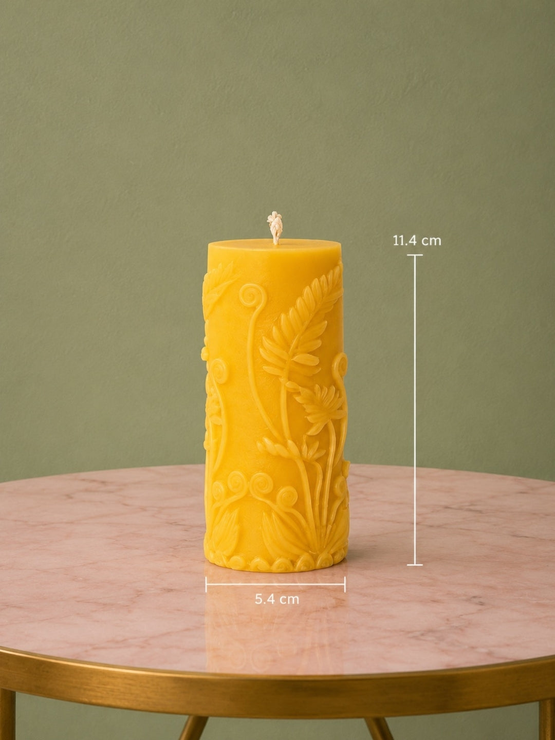 A yellow pillar candle with embossed floral details sits on a pink marble table against a muted green wall. White measurement lines and text indicate a height of 11.
