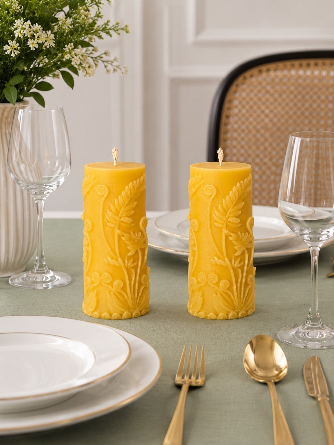 Two yellow pillar candles with embossed floral patterns sit on a green tablecloth in a styled dining setting with white plates, gold cutlery, wine glasses, and a vase of flowers. Velvetine Soirée Beeswax Pillar Candle