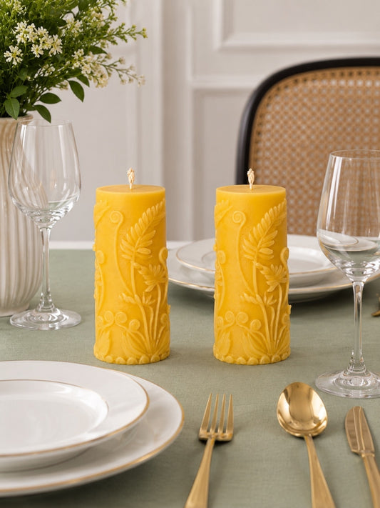 Two yellow pillar candles with embossed floral patterns sit on a green tablecloth in a styled dining setting with white plates, gold cutlery, wine glasses, and a vase of flowers. Velvetine Soirée Beeswax Pillar Candle