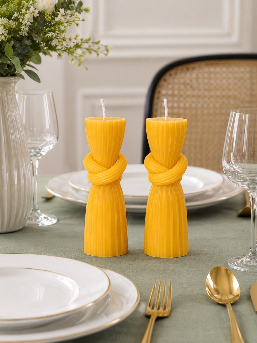 Two bright yellow pleated pillar candles tied in a knot style, displayed on a green tablecloth in a styled dining setting with white plates, gold cutlery, and wine glasses.
