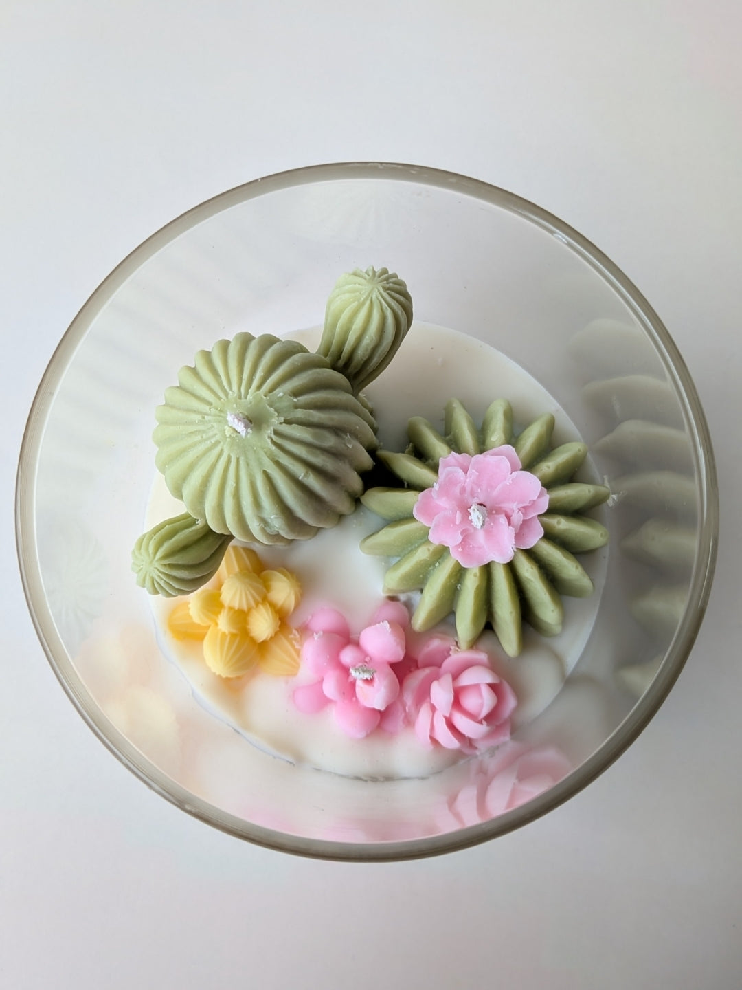 A top-down view of a round glass dish containing a white wax melt or candle decorated with sculpted green succulents, pink flowers, and small yellow accents.