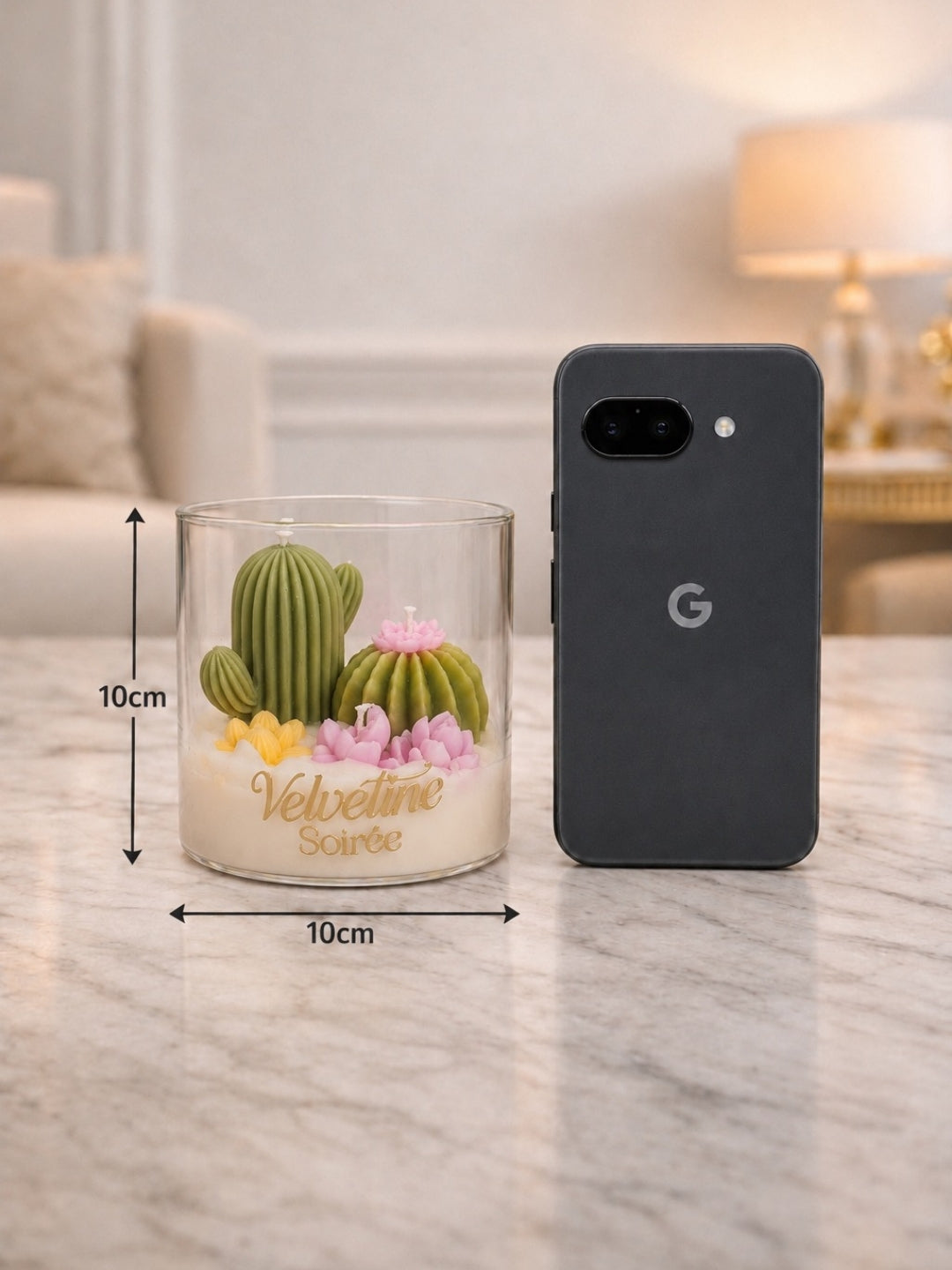 A clear glass candle jar with a cactus and pink flowers, shown next to a dark gray Google Pixel phone case on a marble surface. The image includes dimension lines indicating the jar is 10cm by 10cm.