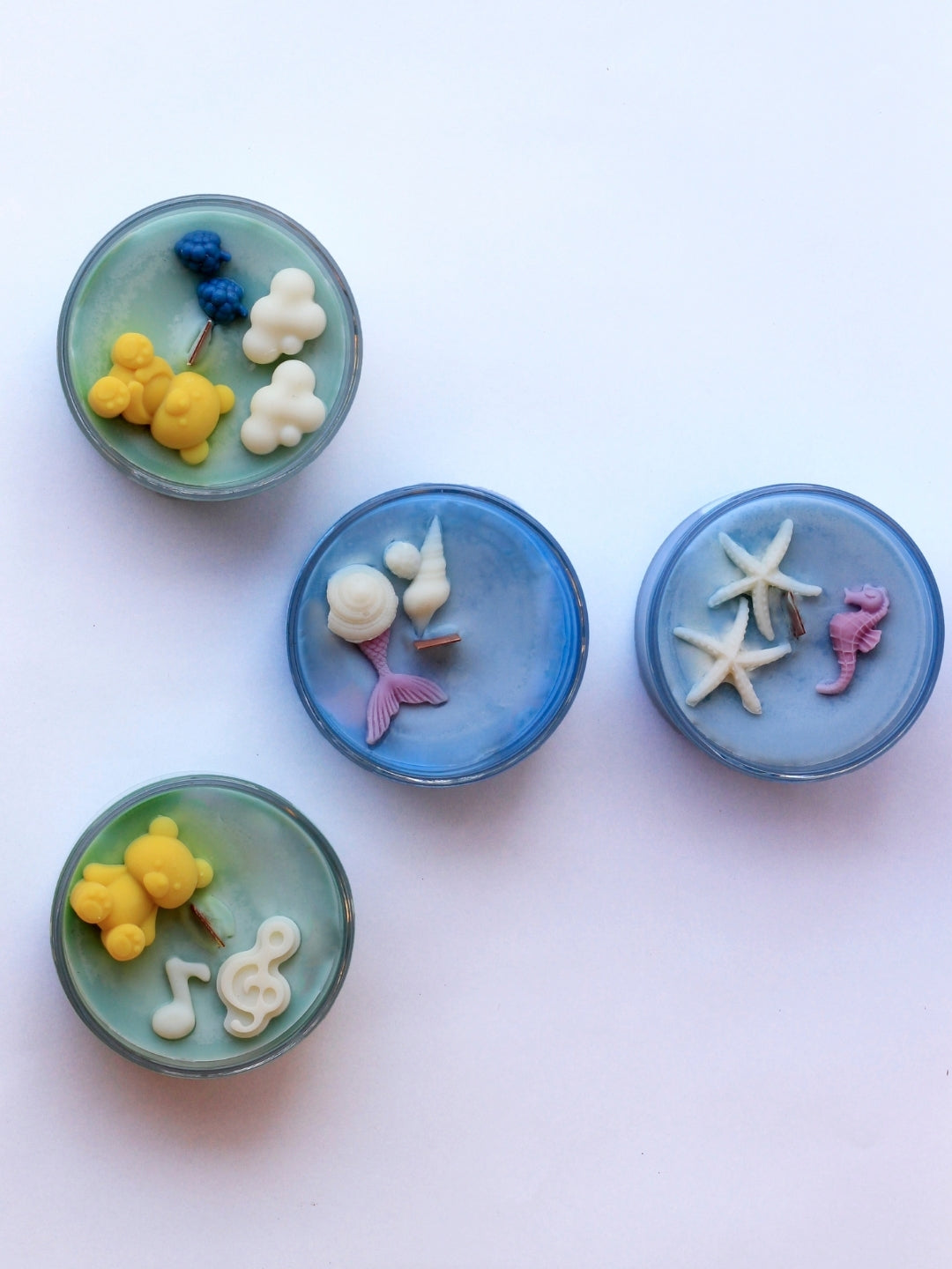 Four Candle Jars  - 2 with sea-themed design and marine fragrance and other 2 with forest design and woody and pine forest fragrance including yellow sea creatures, white clouds, a purple mermaid tail, white starfish, and a pink seahorse, arranged on a light pink background. Velvetine Soirée Candle jars
