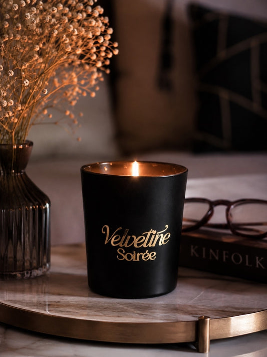 A black scented candle labeled 'Velvetine Soirée' in gold lettering, lit and placed on a marble tray with dried flowers, eyeglasses, and a book in a cozy indoor setting.