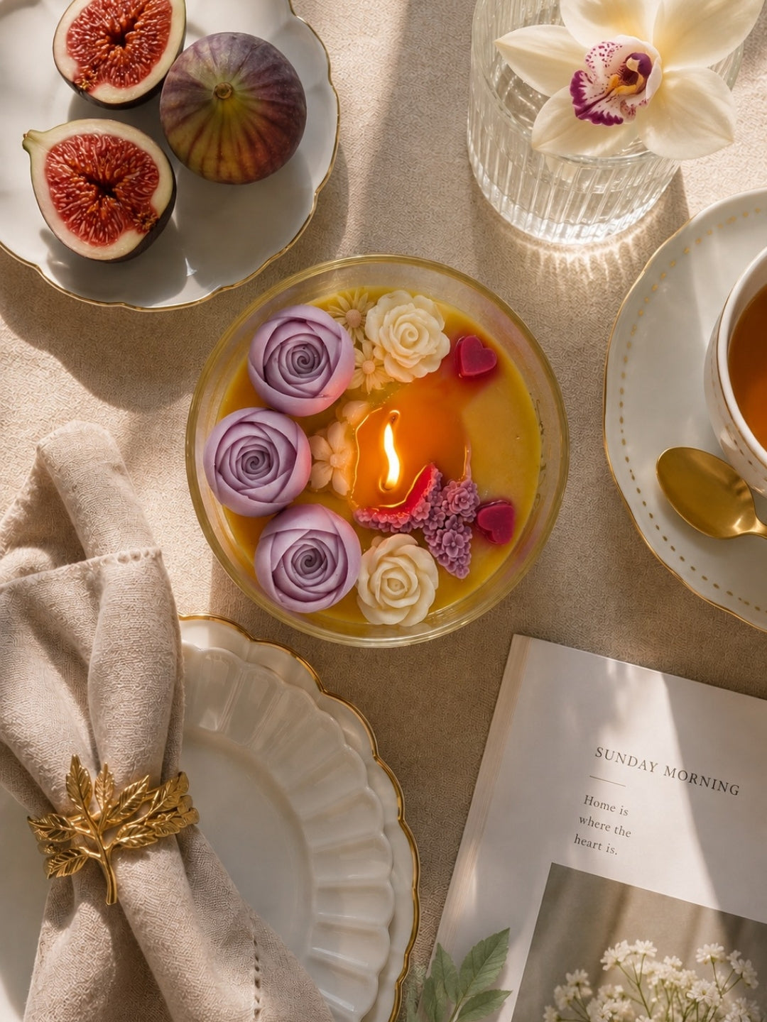 A top-down styled scene around a glass candle jar with a S-Shaped Wooden Wick, decorated with purple and cream flower petals and small pink heart pieces. The setting includes figs on a plate, a white orchid in a glass vase, a cup and saucer with a gold spoon, a folded beige napkin with a gold leaf pin, and a magazine on a beige fabric surface.