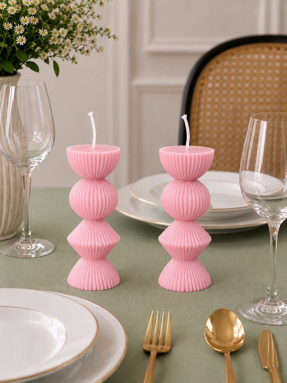 Two pink ribbed pillar candles with white wicks, styled on a green tablecloth with white plates, gold cutlery, wine glasses, and a woven chair in the background. Velvetine Soirée Beeswax Candle Pillar Candles