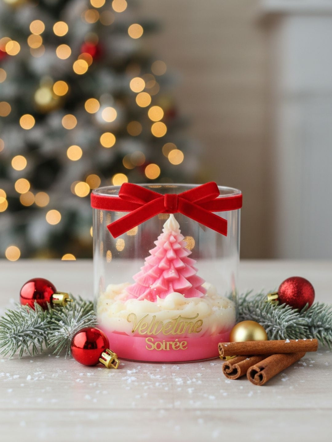 A clear cylindrical glass jar candle with a red ribbon bow around the neck, containing a pink and white layered design that resembles a Christmas tree on top of cream wax. The base of the jar is pink with gold script reading 'Velvetine Soirée'.