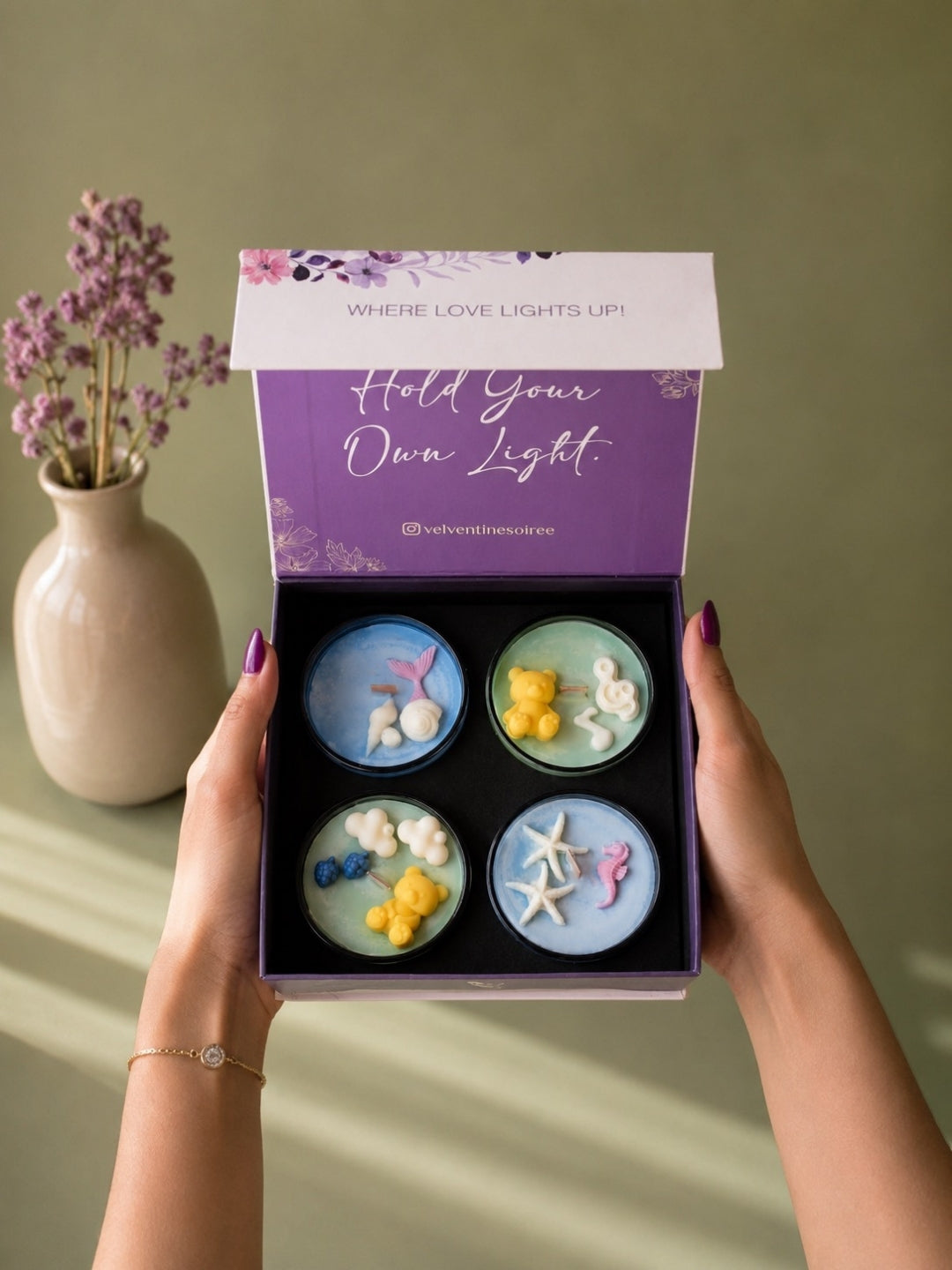A person holds an open gift box containing four round decorative candles with seashell and starfish motifs on a light blue background. The box lid is purple with the text 'Hold Your Own Light!' and a brand handle, while a beige vase with dried flowers sits on a green surface in the background. Marine scented and forest scented candles