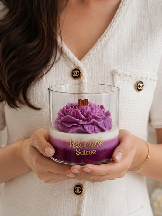 A person in a white textured Chanel-style cardigan holds a clear glass jar candle labeled 'Velvetine Soirée.' The candle has a purple flower-shaped wax top, a white middle band, and a deep purple bottom layer with gold lettering. Sapphire Moonlight Candle Jar with Wooden Wick and Earthy Woody Refreshing Fragrance