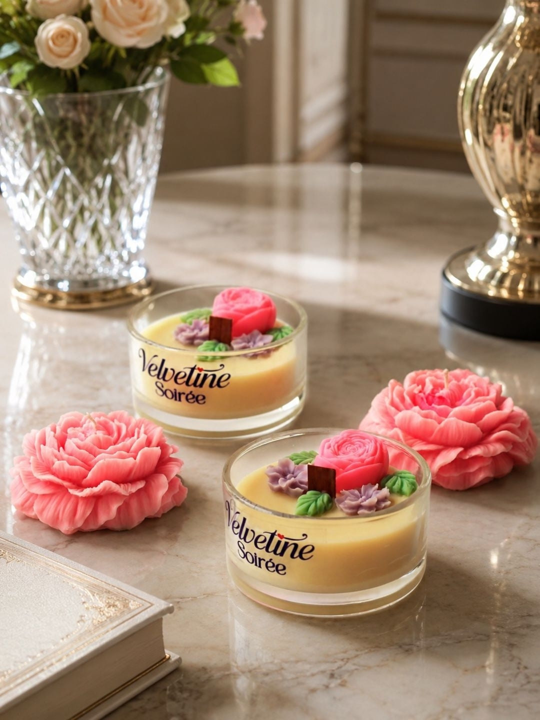 Two small glass jars of Velvetine Soirée candles with Coconut wax and pink roses decorations, styled on a marble tabletop with pink peony flowers and