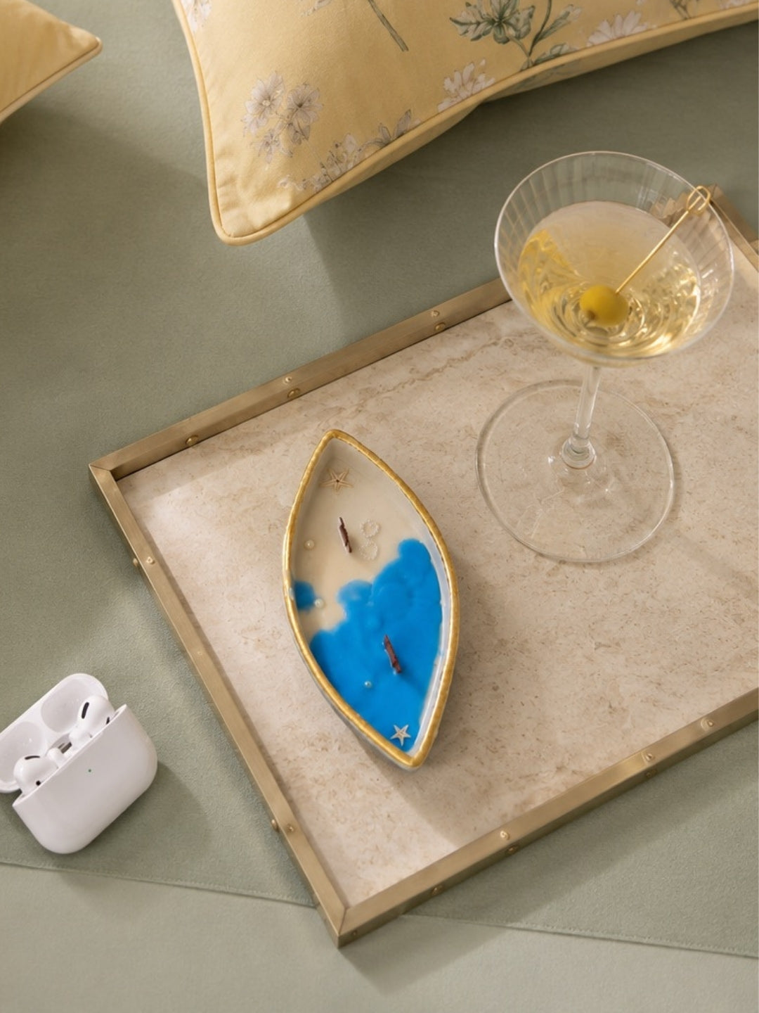A decorative boat-shaped jar with a blue and white coloured coconut wax  candle with 2 wooden wicks and gold trim, styled on a beige tray with a cocktail glass, earbuds case, and yellow floral pillows on a green surface.