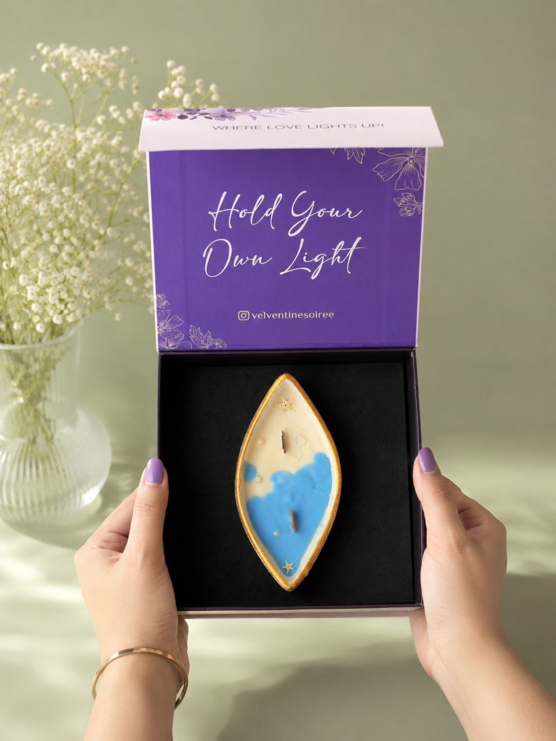 A person holds an open gift box containing a decorative boat-shaped candle with a blue and cream coconut wax and  2 wooden wicks  and gold trim. The box lid is purple with the text 'Hold Your Own Light' and the brand name 'Velvetine Soirée;