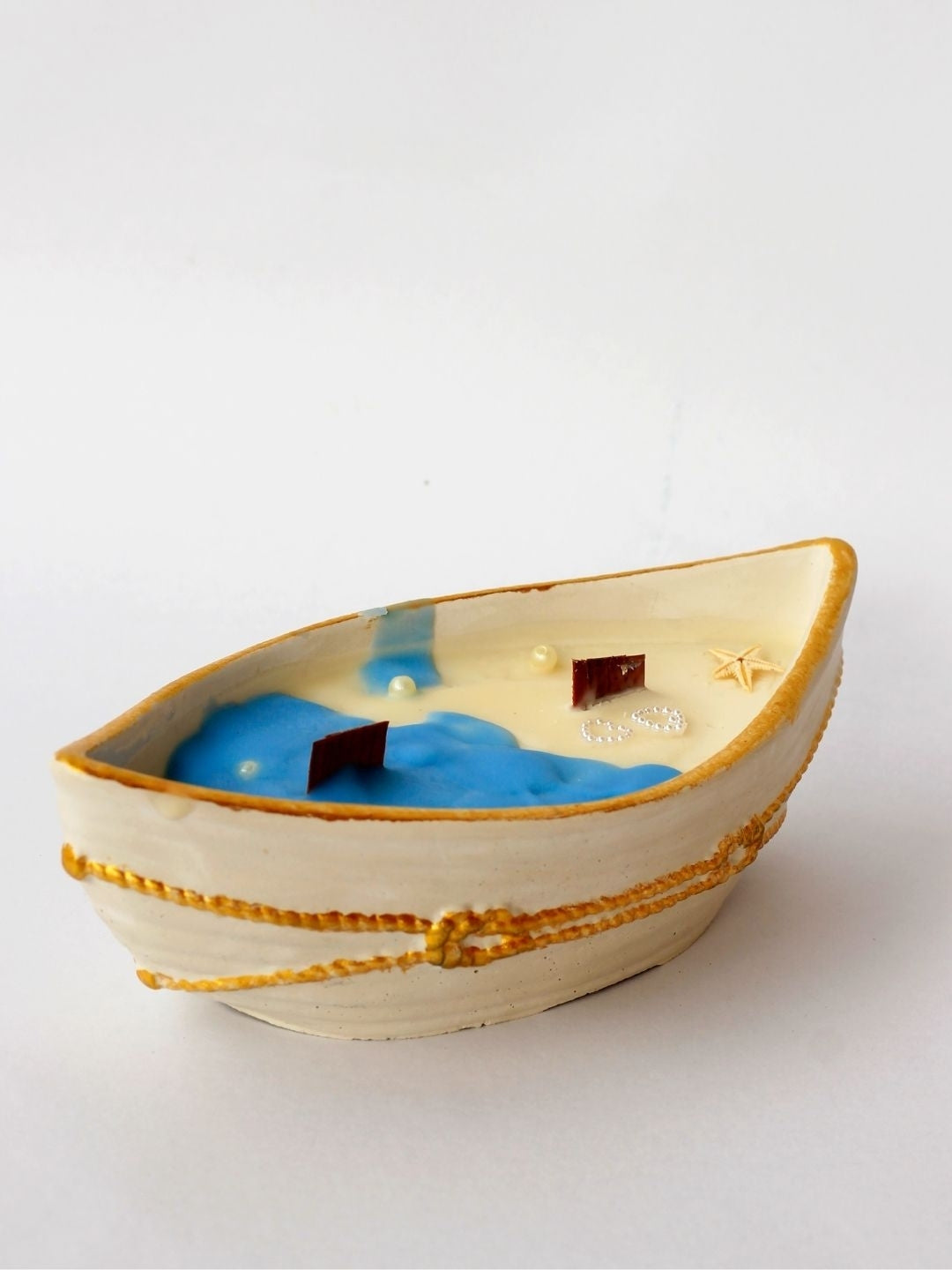 A small boat-shaped decorative candle with a cream-colored exterior, gold rope details, and a blue ocean coconut wax with small starfish and 2 wooden wicks, shown on a plain white background.
