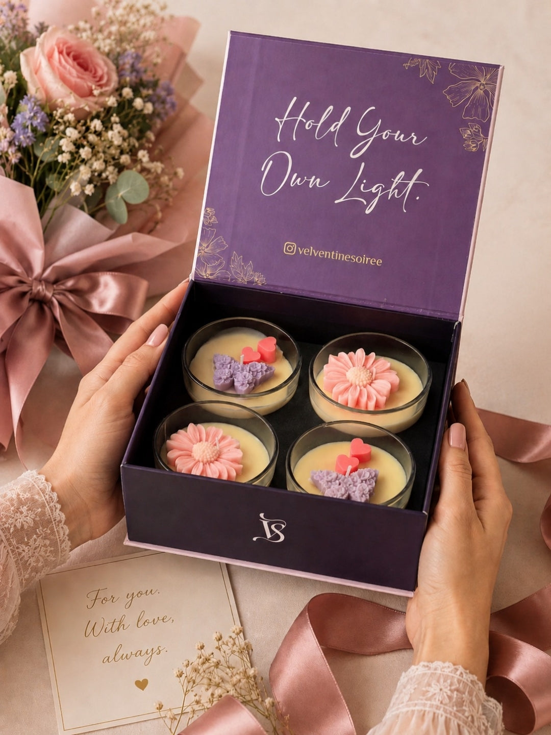 A person holds an open purple gift box containing four Velvetine Soiree Candle jars filled with coconut wax and pink and purple flower-shaped toppers. The box lid reads 'Hold Your Own Light' with the brand name "Velvetine Soirée" visible below.